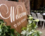 Drewniana tablica "Welcome to Our Wedding" ozdobiona zielenią. W tle widać przygotowane stoły i krzesła, a także obsługę. Opłata ZAIKS za muzykę na weselu.
