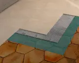 Dettaglio di un pavimento in fase di posa: nuove piastrelle beige sopra uno strato di colla, membrana impermeabilizzante turchese e vecchie piastrelle esagonali color terra.