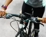 Ręce na kierownicy roweru, gotowe do jazdy. Widać manetki, hamulce i uchwyt na telefon. Trwa montaż licznika rowerowego.