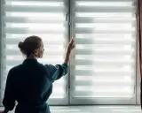 Une femme ajuste un store enrouleur blanc, laissant entrer la lumière du jour.