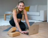 Glad kvinna förbereder sig för ett Anusara yoga-pass hemma, med en laptop och yogablock redo.