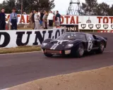 Ein schwarzer Ford GT40 Mk3 mit der Startnummer 2 rast auf der Rennstrecke, während Zuschauer und Werbetafeln im Hintergrund zu sehen sind.