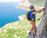 Mężczyzna na via ferrata Krucze Skały, zmagający się ze stromą ścianą skalną, z widokiem na błękitne morze i zielone wybrzeże.