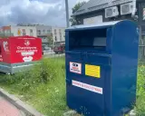 Kontener na odzież używaną obok czerwonego przyczepy z napisem "Charytatywna zbiórka odzieży". To miejsce, gdzie można oddać tekstylia, odpady ubraniowe, zamiast wyrzucać.