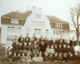 Schulkinder und Lehrer posieren vor einem Gebäude. Die Kleidung deutet auf eine Zeit hin, in der Armut, also Pauperismus, ein großes Thema war.