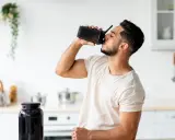Mann trinkt Proteinshake aus schwarzem Shaker. Daneben steht ein großer Behälter mit Pulver. Wozu Protein**pulver**? Für Muskelaufbau und Erholung.