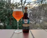 Szklanka z bursztynowym piwem Grolsch Royale Weizen IPA i butelka obok, na tle drzew i nieba.