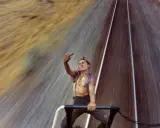 Mike Brodie, un giovane ribelle, fa il gesto del dito medio mentre viaggia su un treno merci, sfidando il mondo con uno sguardo provocatorio.