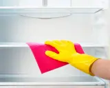 Mano con guante amarillo limpiando estante de frigorífico vacío con paño rosa.