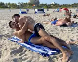 Two men embrace on a towel at Haulover Beach, a popular spot for gay beachgoers. Other sunbathers relax on the sand in the background.