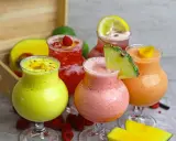 Cinq verres de boisson énergisante maison aux fruits colorés : jaune, rose, rouge et orange, garnie de fruits frais.