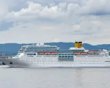 La nave da crociera Costa Romantica naviga in acque calme, con montagne verdi sullo sfondo e un cielo nuvoloso.