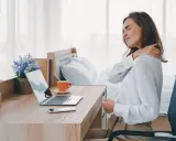 Femme se massant l'épaule, assise à son bureau. La mauvaise position bureau peut causer des douleurs.