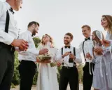 Pan młody i świadek świętują z panną młodą i druhnami. Wszyscy trzymają kieliszki, a jeden z panów otwiera butelkę szampana.