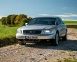 Silberner Audi A8 D2 W12 auf Schotterweg, umgeben von grünen Feldern und Bäumen im Herbstlicht.