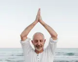 Mężczyzna z zamkniętymi oczami, ręce złożone nad głową, praktykuje mindfulness co to. Spokój nad morzem.