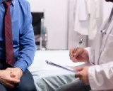 Mężczyzna w niebieskiej koszuli i krawacie siedzi na kozetce lekarskiej, rozmawiając z lekarzem o słabym strumieniu moczu.