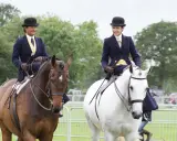 Zwei Reiterinnen im Damensattel, elegant gekleidet, reiten auf braunen und weißen Pferden.