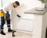 Un technicien en bleu, avec une ceinture à outils, installe un radiateur à inertie. Il est prêt à brancher le radiateur inertie sur prise de courant.