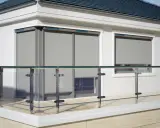 Nowoczesne okno przy samej ścianie z szarymi roletami i szklaną balustradą na balkonie.