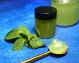Une cuillère dorée présente une portion de gelée de menthe fraîche. À côté, un pot fermé et un autre ouvert contiennent cette même gelée verte et rafraîchissante.
