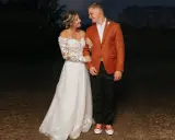 A bride in a lace gown and groom in a rust-colored jacket and black pants, sporting red sneakers, showcase their semi-formal attire for wedding.