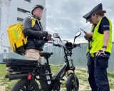 Dostawca z żółtą torbą na plecach rozmawia z policjantkami obok swojego roweru elektrycznego bez pedałowania.