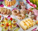 Kolorowy stół z pysznym śniadaniem wielkanocnym: babka, kiełbaski, sałatka, babeczki, sernik i jajka faszerowane.