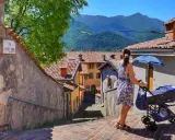 Una donna spinge un passeggino con neonati in montagna, tra case con tetti rossi e un panorama mozzafiato.
