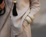 Dettaglio di un outfit in lino materiale, con una mano che tiene una borsa nera, un ciondolo con nappa nera e bracciali color crema.