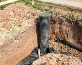 Wykop pod budowę, w którym widać połączenie rury drenarskiej z PCV z zieloną pokrywą.