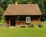 Drewniana chata z glinianym dachem, typowa dla skansenów w Polsce, otoczona zielenią i drewnianym płotem.