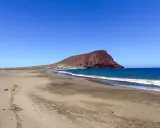 Plaże z białym piaskiem na Teneryfie, z charakterystyczną czerwoną górą w tle i turkusowym oceanem.