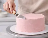 Manos expertas aplican glaseado rosa a un pastel redondo. Aprende como decorar tartas con esta técnica.