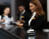 Dzwonek przy ladzie hotelowej, gdzie pracownicy obsługują gości. Stanowiska pracy w hotelu tętnią życiem.
