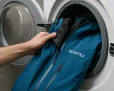 Mano mete chaqueta azul GORE-TEX en lavadora. Aprende cómo lavar Goretex para mantener su impermeabilidad.