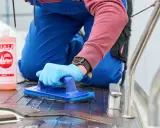 Person reinigt mit blauer Bürste und SEMCO Teak Cleaner das Holzdeck eines Bootes.