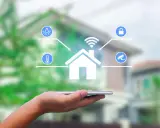 Dłoń trzyma smartfon, z którego wyłania się symbol domu z ikonami oświetlenia, temperatury, zamka i kamery. To wizualizacja systemu zarządzania energią.