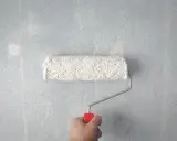 Main tenant, comment rattraper une peinture mal faite. Un rouleau à peinture blanc est tenu par une main sur un mur bleu clair texturé.