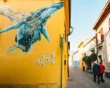 Un murale di balene blu su un muro giallo a San Giuliano Rimini. Una coppia passeggia lungo la strada acciottolata.