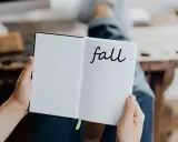 Otwarta książka z napisem "fall" na prawej stronie. Osoba w dżinsach trzyma ją, przygotowując się do jesiennych zapisków.