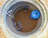 Wnętrze studni głębinowej z niebieskim zbiornikiem hydroforowym i systemem rur. Widać manometr i zawory.