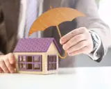 Main d'un homme tenant un parapluie en bois au-dessus d'une maison miniature. Une image symbolisant l'assurance habitation obligatoire.