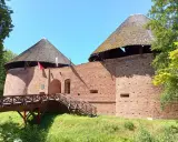 Zamek międzyrzecz z drewnianym mostem i wieżami o stożkowych dachach. Czerwona cegła i zieleń otoczenia.