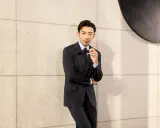 A man in a black suit and black shirt, holding sunglasses, leans against a textured wall.