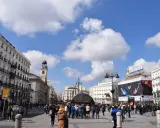 Menschenmassen beleben den Platz Puerta del Sol in Madrid unter einem blauen Himmel mit Wolken. Der Uhrturm und das Tio Pepe Schild sind sichtbar.