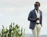 Man in stylish beach wedding attire, holding a drink, with cacti and a hazy sky.