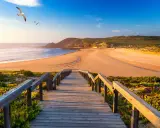 Holzsteg führt zu einem goldenen Strand in Aljezur, Portugal. Möwen kreisen über dem Meer unter blauem Himmel.
