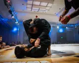 Dwóch mężczyzn w czarnych kimonach walczy w parterze. Intensywna walka jiu jitsu, z widownią w tle.
