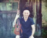 Uśmiechnięty mężczyzna w kapeluszu trzyma ukulele, gotowy do grania nim zajdzie słońce.